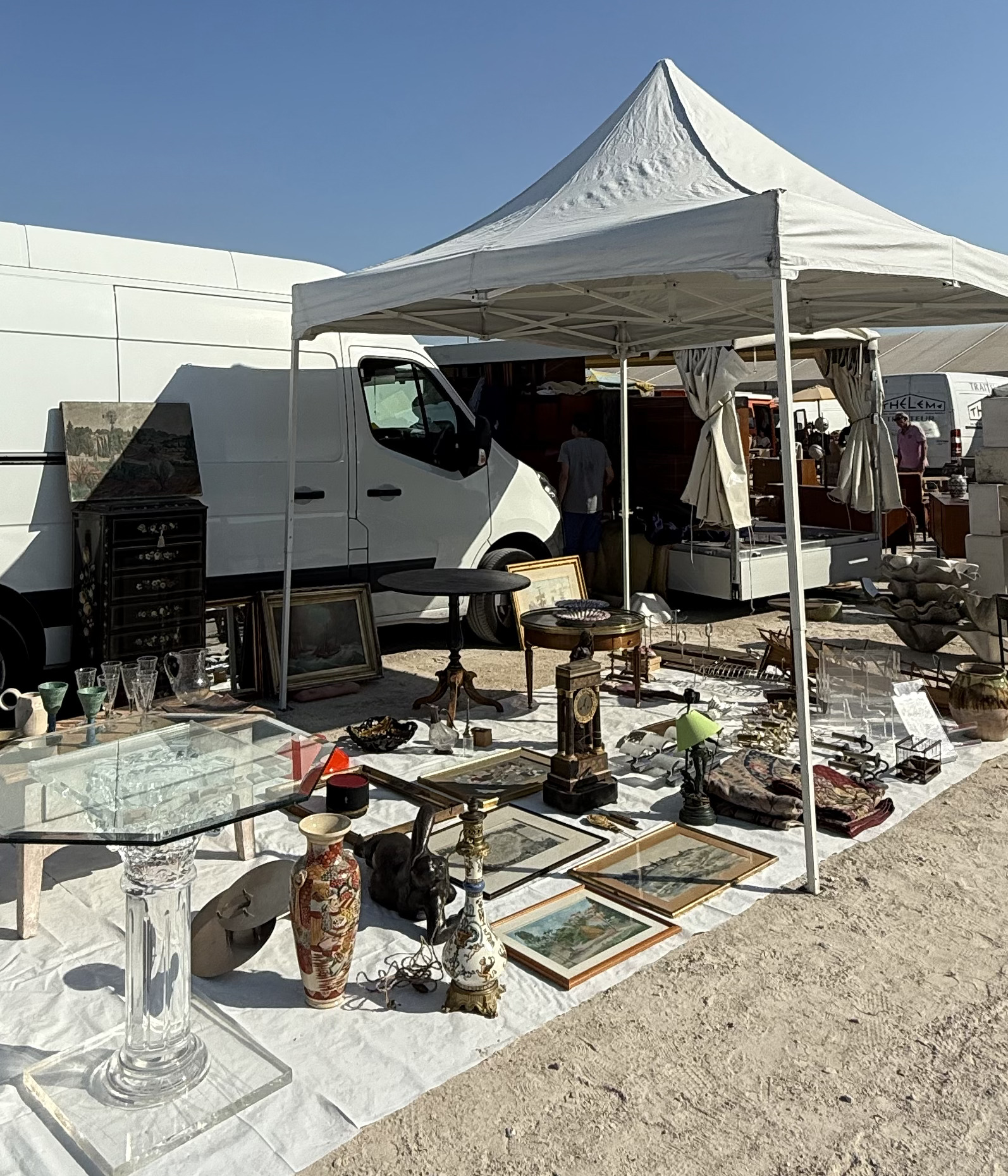 Stand de brocante sous barnum : table à plateau de verre, tableaux encadrés, vases et bronzes